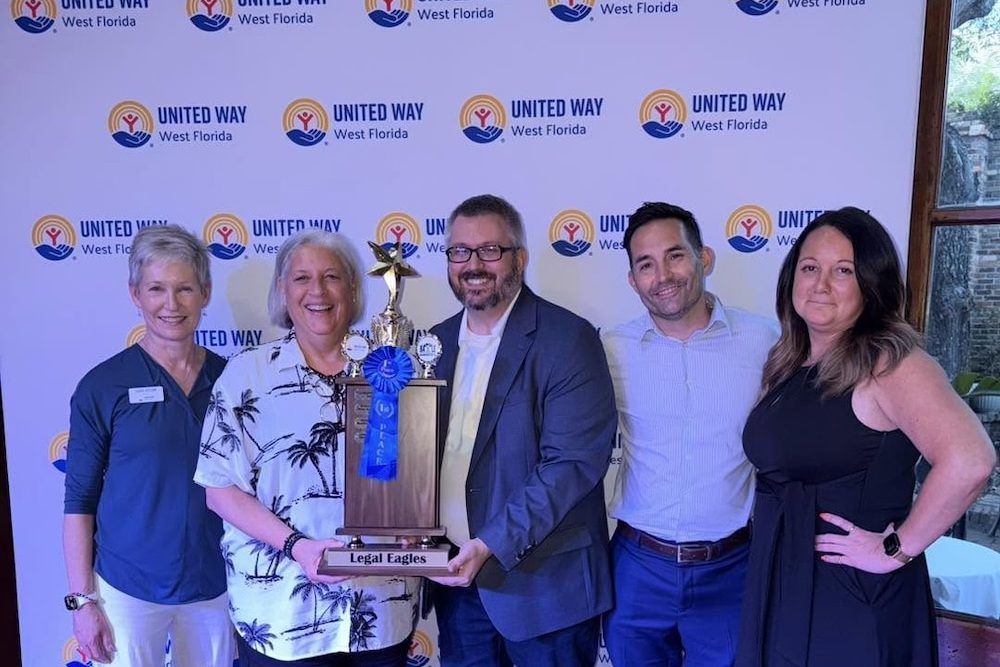 Five adults pose with a large trophy reading 'Legal Eagles' in front of a United Way West Florida backdrop.