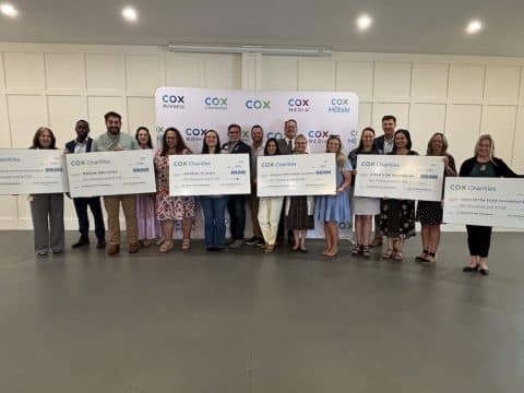 Group of people standing in a line, each holding a large ceremonial check, in front of a Cox Charities backdrop.