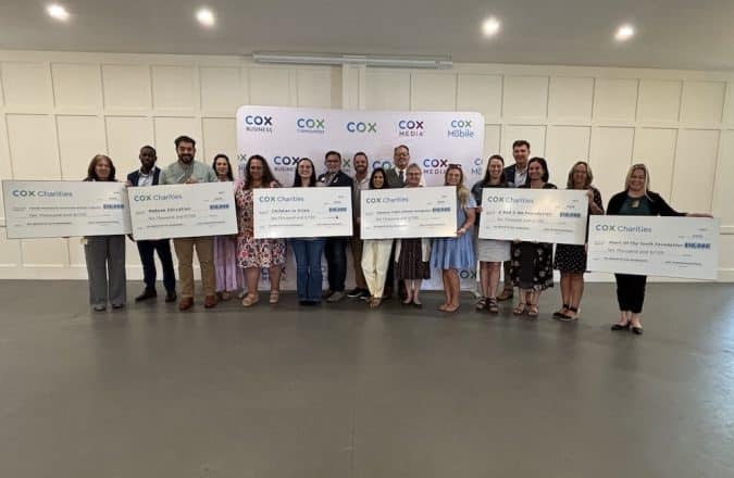 Group of people standing in a line, each holding a large ceremonial check, in front of a Cox Charities backdrop.