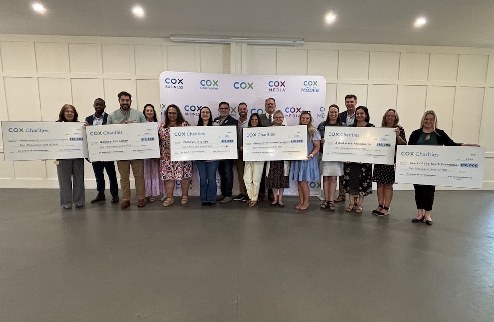Group of people standing in a line, each holding a large ceremonial check, in front of a Cox Charities backdrop.