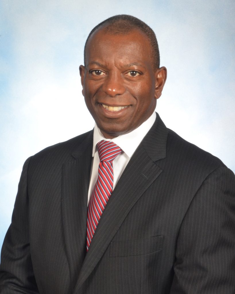Professional headshot of a smiling man in a dark suit and red striped tie, against a light blue background.