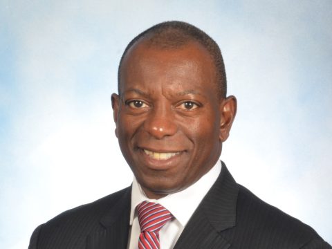 Professional headshot of a smiling man in a dark suit and red striped tie, against a light blue background.