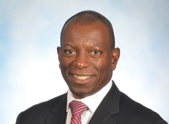 Professional headshot of a smiling man in a dark suit and red striped tie, against a light blue background.