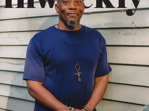 Portrait of a smiling man in a blue shirt and beige cap, wearing a pendant necklace and beaded bracelets, standing against a pale wooden wall with large 'weekly' text across the top.