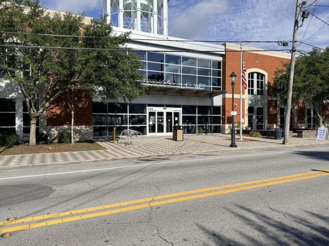 Public civic building with a glass entrance, brick facade, and an American flag outside on a sunny day.