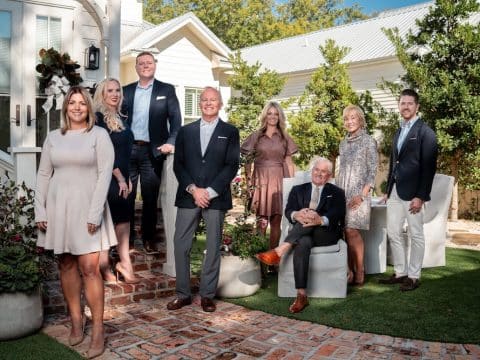 Group portrait of eight adults in business attire posing outside a white house with brick steps and potted plants on a sunny day.