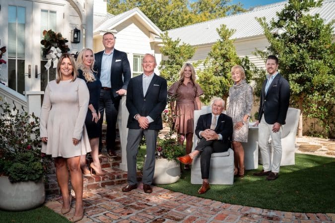 Group portrait of eight adults in business attire posing outside a white house with brick steps and potted plants on a sunny day.
