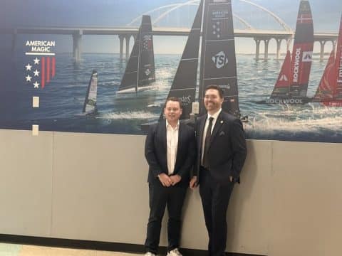 Two men in suits pose for a photo in front of a sailboat racing mural at an airport or similar venue, with American Magic branding in the background.