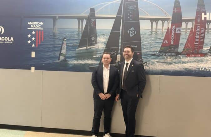 Two men in suits pose for a photo in front of a sailboat racing mural at an airport or similar venue, with American Magic branding in the background.