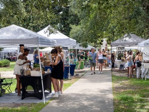 Outdoor market with white canopy tents and people browsing vendor booths along a paved path in a sunny park area