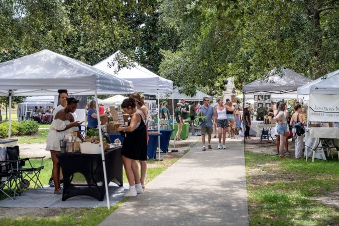 Outdoor market with white canopy tents and people browsing vendor booths along a paved path in a sunny park area