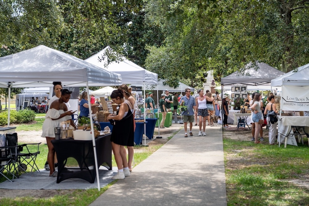Outdoor market with white canopy tents and people browsing vendor booths along a paved path in a sunny park area