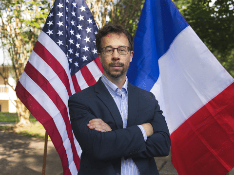 Man in a navy blazer and glasses stands with arms crossed, flanked by the American and French flags outdoors.