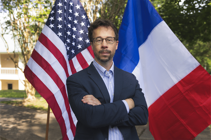 Man in a navy blazer and glasses stands with arms crossed, flanked by the American and French flags outdoors.