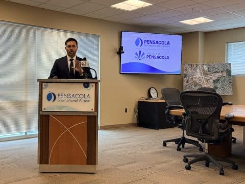Male presenter in a suit at a podium in a conference room, Pensacola International Airport signage visible on the podium and a wall screen behind him displaying the Pensacola logos.