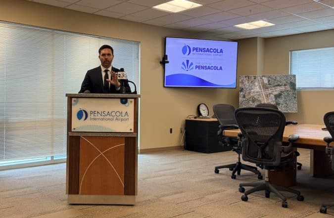 Male presenter in a suit at a podium in a conference room, Pensacola International Airport signage visible on the podium and a wall screen behind him displaying the Pensacola logos.