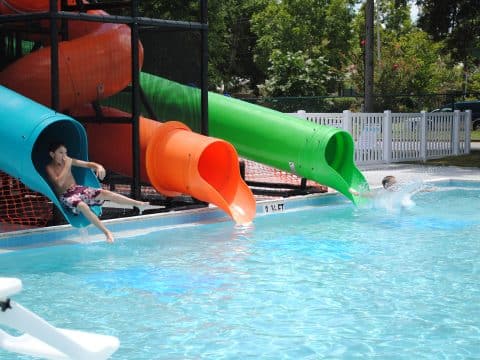 Boy exiting a blue water-slide tube into a pool under colorful tubes at a water park, with orange and green slides nearby.