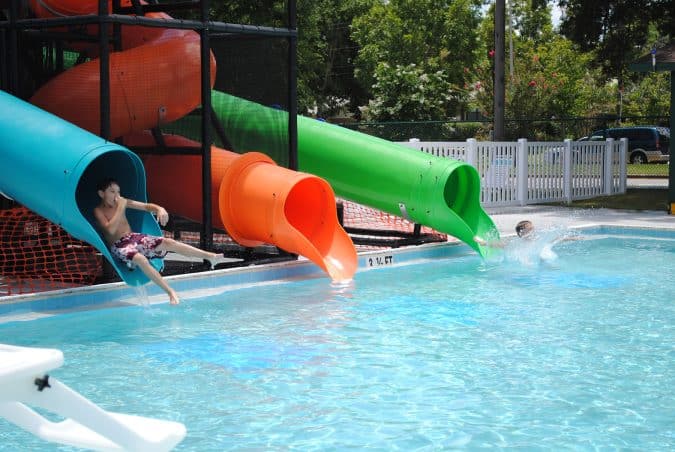 Boy exiting a blue water-slide tube into a pool under colorful tubes at a water park, with orange and green slides nearby.
