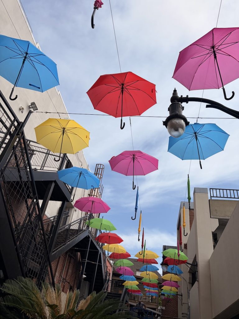 Colorful umbrellas hung overhead across a narrow street between buildings, creating a bright canopy against a blue sky.