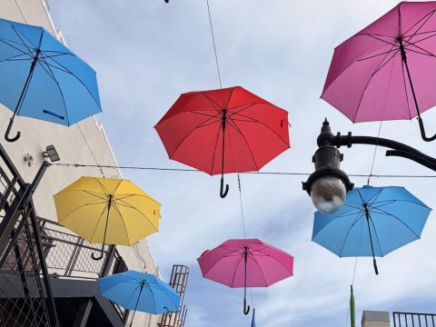 Colorful umbrellas hung overhead across a narrow street between buildings, creating a bright canopy against a blue sky.