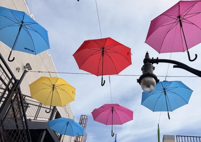 Colorful umbrellas hung overhead across a narrow street between buildings, creating a bright canopy against a blue sky.