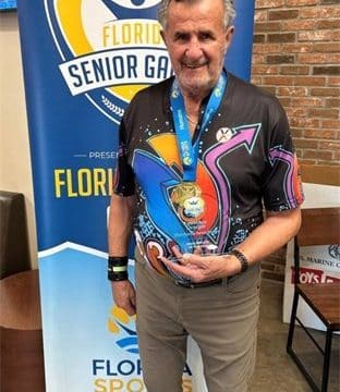 Smiling older man wearing a vibrant shirt, holding a gold medal, standing in front of a Florida Senior Games banner.
