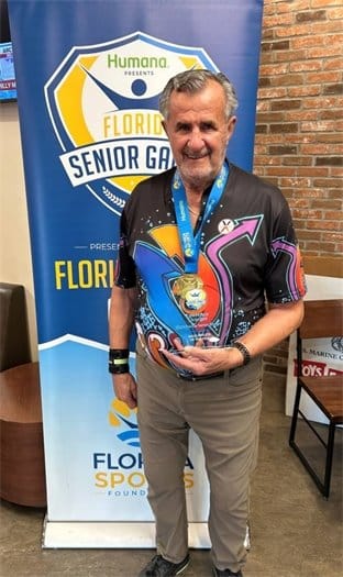 Smiling older man wearing a vibrant shirt, holding a gold medal, standing in front of a Florida Senior Games banner.
