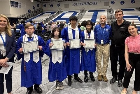 Group of five students in blue graduation gowns holding certificates, flanked by staff in a gym with a 'Go Wildcats' banner in the background.
