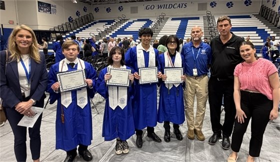 Group of five students in blue graduation gowns holding certificates, flanked by staff in a gym with a 'Go Wildcats' banner in the background.