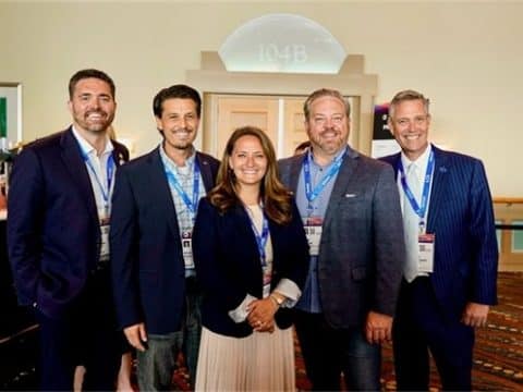 Five professionally dressed individuals posing for a group photo at a conference, all wearing lanyards with badges, in a hotel lobby setting.