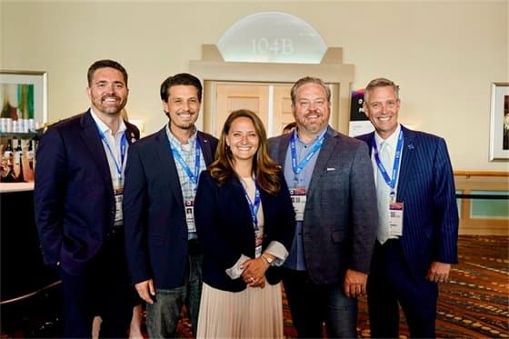 Five professionally dressed individuals posing for a group photo at a conference, all wearing lanyards with badges, in a hotel lobby setting.