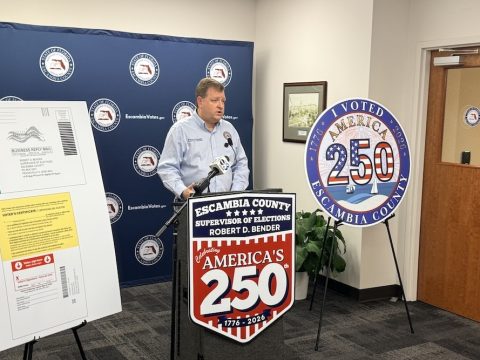 Man at a podium during an Escambia County election event, with a large 'America's 250' sign on display behind him.