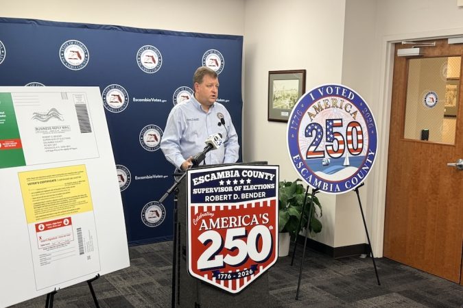 Man at a podium during an Escambia County election event, with a large 'America's 250' sign on display behind him.