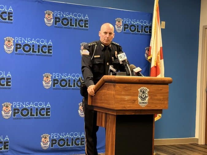 Bald police officer in uniform speaking at a wooden podium with Pensacola Police backdrop behind him.