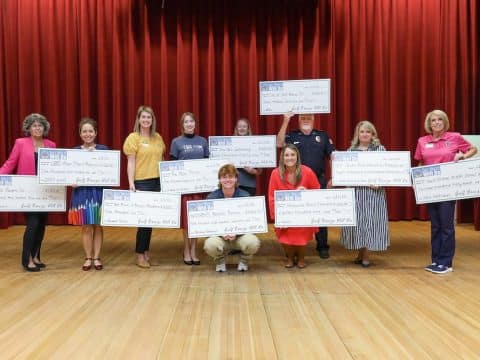 Group of volunteers on a stage holding oversized ceremonial checks during a grant/award ceremony with a red curtain backdrop.