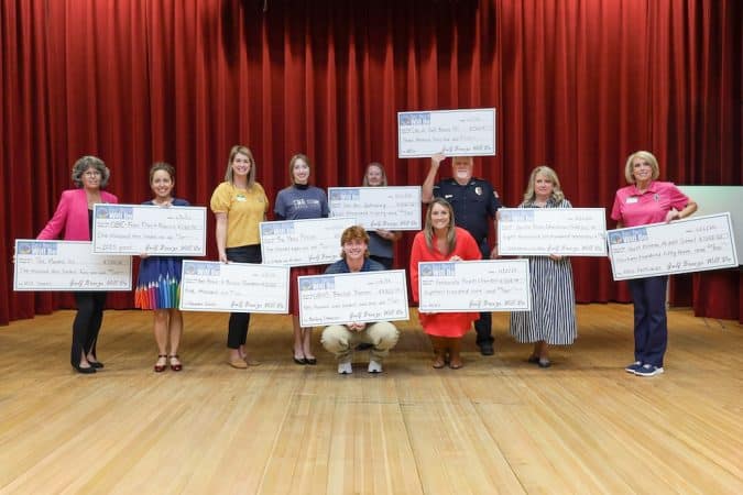Group of volunteers on a stage holding oversized ceremonial checks during a grant/award ceremony with a red curtain backdrop.