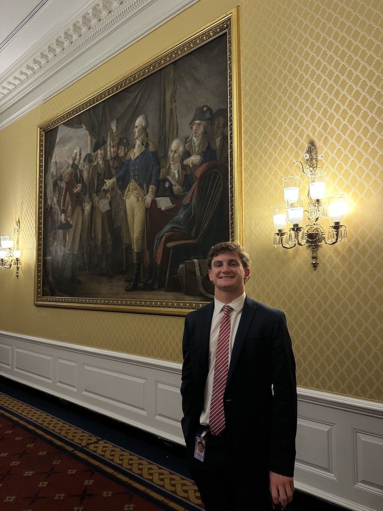 A young man in a dark suit and striped tie stands in a gilded hallway beside a large framed historical painting.
