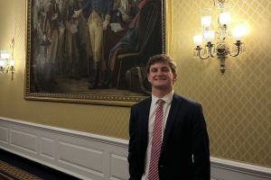 A young man in a dark suit and striped tie stands in a gilded hallway beside a large framed historical painting.