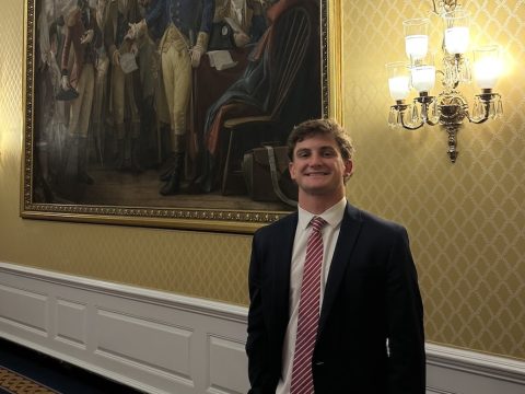 A young man in a dark suit and striped tie stands in a gilded hallway beside a large framed historical painting.