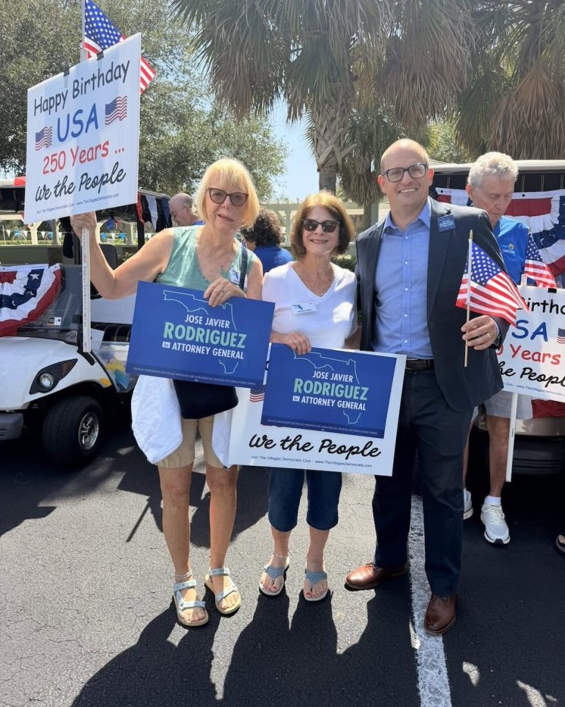 Group of four adults at a political event holding blue 'Jose Javier Rodriguez for Attorney General' signs, with American flags and a 'Happy Birthday USA' sign in the background.