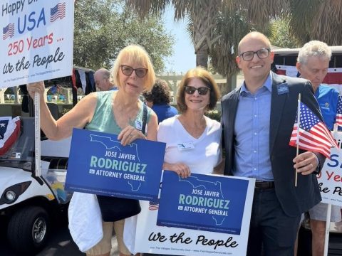 Group of four adults at a political event holding blue 'Jose Javier Rodriguez for Attorney General' signs, with American flags and a 'Happy Birthday USA' sign in the background.