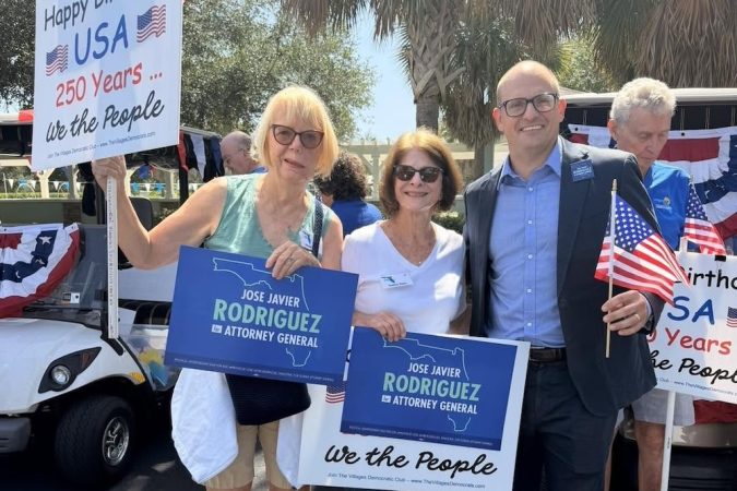Group of four adults at a political event holding blue 'Jose Javier Rodriguez for Attorney General' signs, with American flags and a 'Happy Birthday USA' sign in the background.