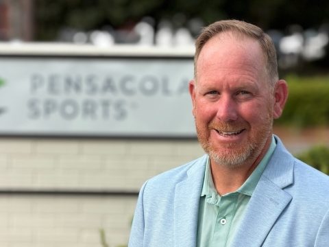 Smiling man in a light blue blazer standing outdoors in front of a Pensacola Sports sign.