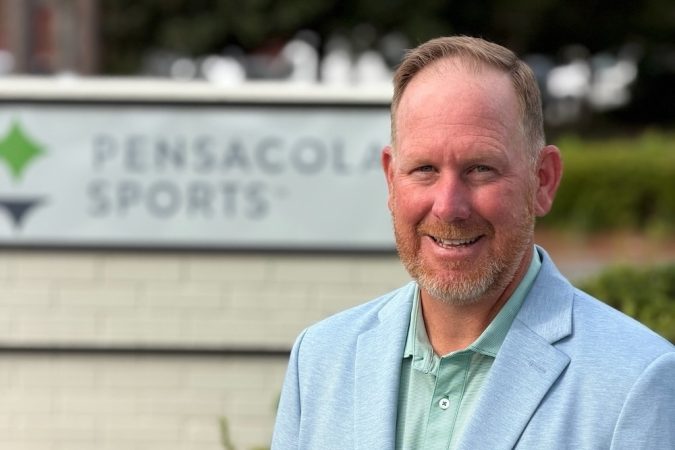 Smiling man in a light blue blazer standing outdoors in front of a Pensacola Sports sign.