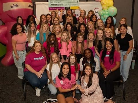 Group of smiling young women posing for a photo at an indoor event, many wearing pink shirts and casual outfits, with balloons in the background.