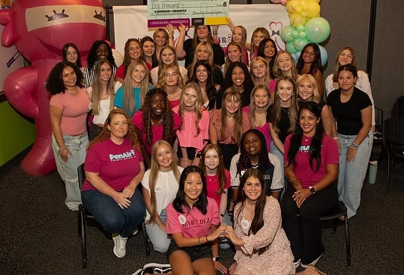 Group of smiling young women posing for a photo at an indoor event, many wearing pink shirts and casual outfits, with balloons in the background.