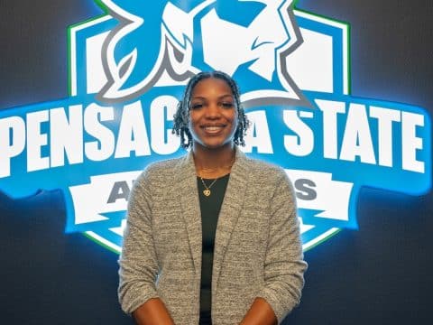 Smiling woman in a gray cardigan posing in front of a blue Pensacola State Athletics logo on a dark wall.