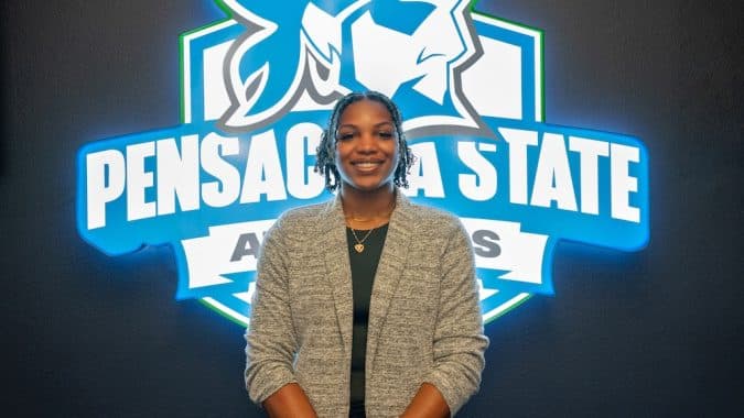 Smiling woman in a gray cardigan posing in front of a blue Pensacola State Athletics logo on a dark wall.