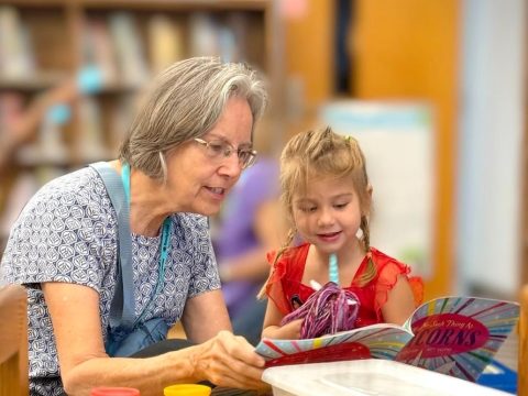 Older woman and young girl reading a colorful book together at a crafts table in a library.