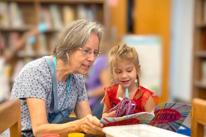 Older woman and young girl reading a colorful book together at a crafts table in a library.
